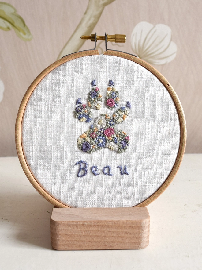 Floral Meadow Dog Paw, of Lavender Blues and Buttermilk yellow blossoms.  Displayed in an embroidery hoop on a wooden stand.
