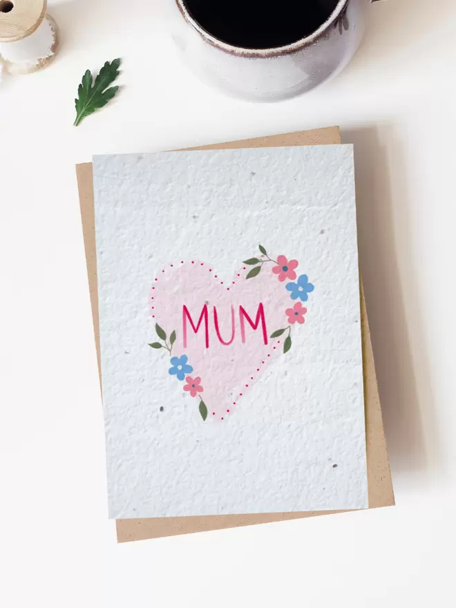 Seeded paper greeting card featuring a heart illustration with flowers with 'Mum' in the centre. Placed on a white background with a cup of coffee and a leaf at the top of the image,