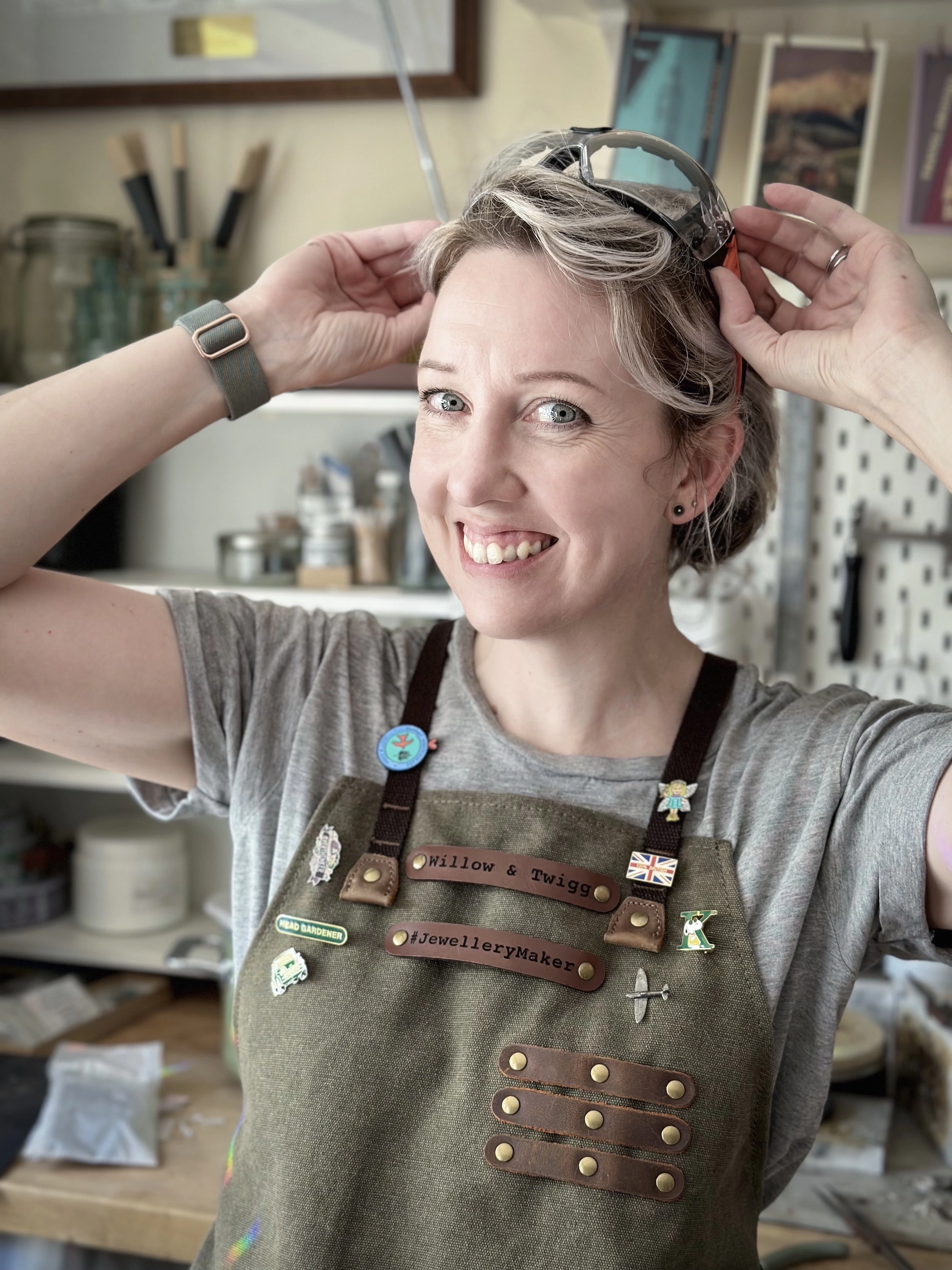 Kelly from Willow And Twigg, standing in her jewellery studio wearing a green canvas apron and placing a pair of safety goggles on her head.