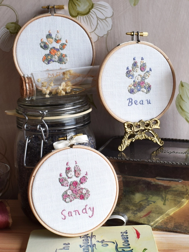 A display of 3 Dog Paw embroidery kits in their hoops, top to bottom are 1. Sunshine Garden (Oranges, Yellows, Purples).  2.  Floral Meadow (Lavender Blues, Magenta & Buttermilk Yellow).  3.. Pink Roses (5 shades of pink).