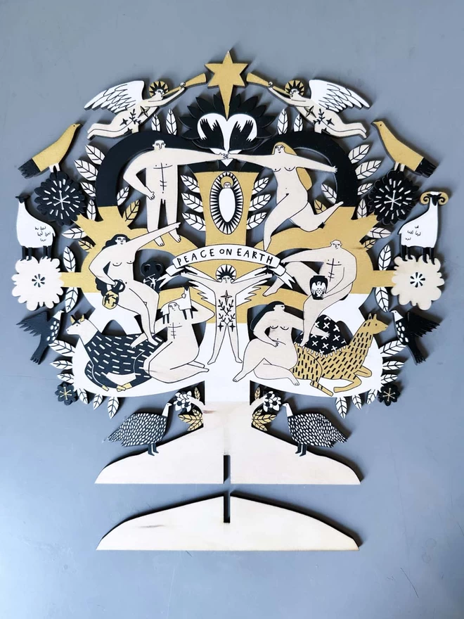 A wooden laser cut nativity, printed in black, white and gold is laid flat with its stand