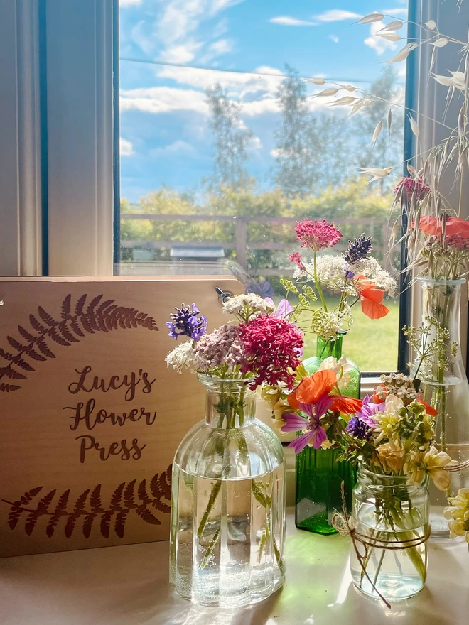 Lucy’s Studio Window Sill with Jars and Bottles Holding Flowers Ready for Pressing; Lucy’s Flower Press in the Background