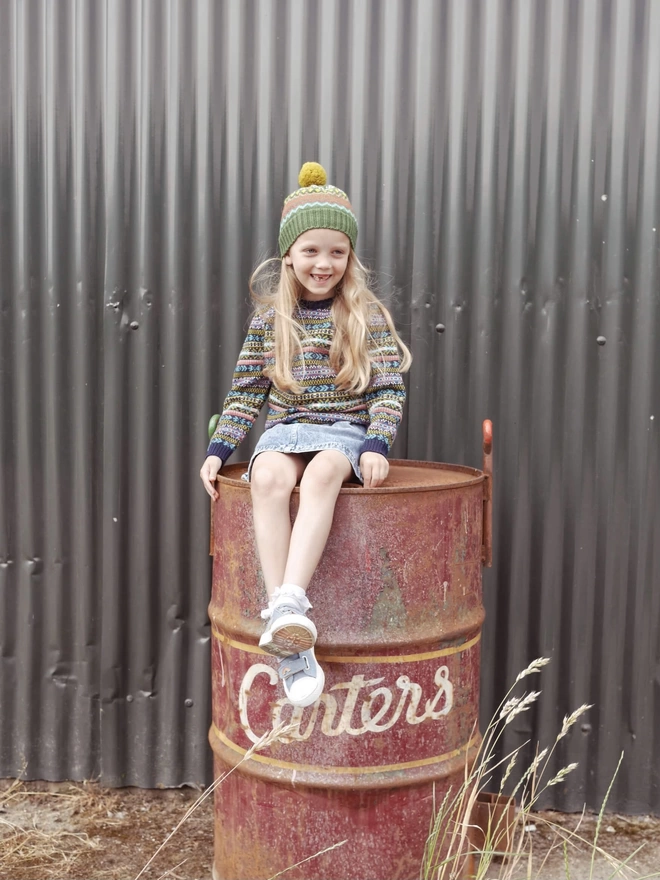Girl sitting on an oil drum wearing the Voyager knitted hat.