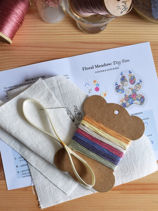 Embroidery Kit contents displayed on top of a pine chest.  Showing: thread card with coloured yarns, pre-printed design on linen, a length of ribbon, a needle, an extra linen and full instructions.