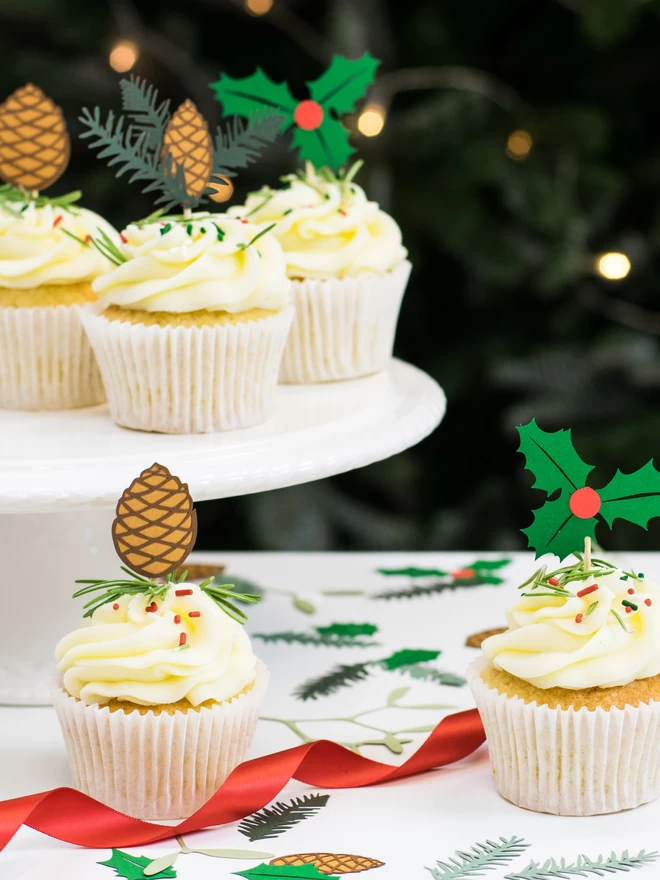6 cupcakes, each decorated with a paper holly, spruce or pinecone cake decoration are displayed on a table decorated for Christmas
