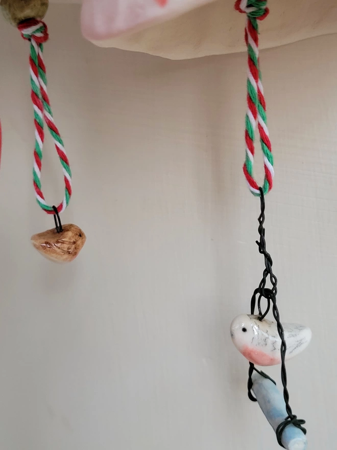 close up of a ceramic robin bird  bead strung on a coloured twine from a ceramic bell there is a pink budgie bead hanging over a swing  also stung on colourful string