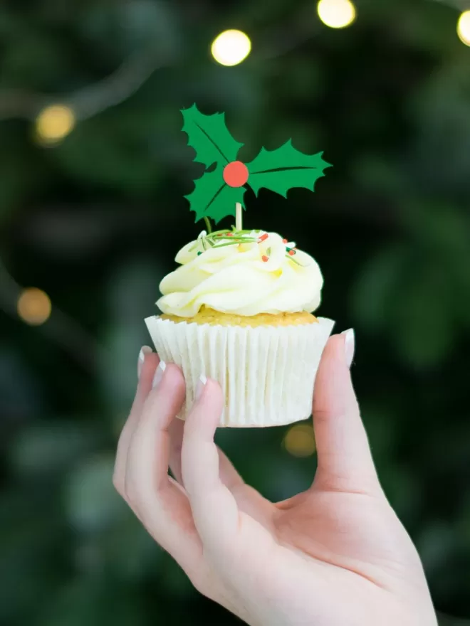 A christmas cupcake held in hand.  Paper cake topper is a sprig of holly with three leaves and a single central red berry