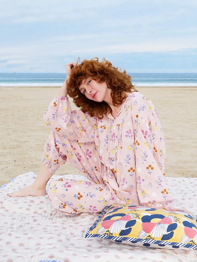 red hair girl wearing a pale pink block printed pyjama in pastel colours on a beach and holding a bright yellow cushion
