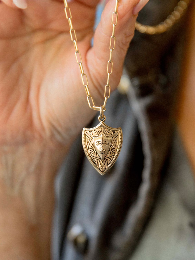 Gold Joan of Arc shield necklace hanging from women's hand