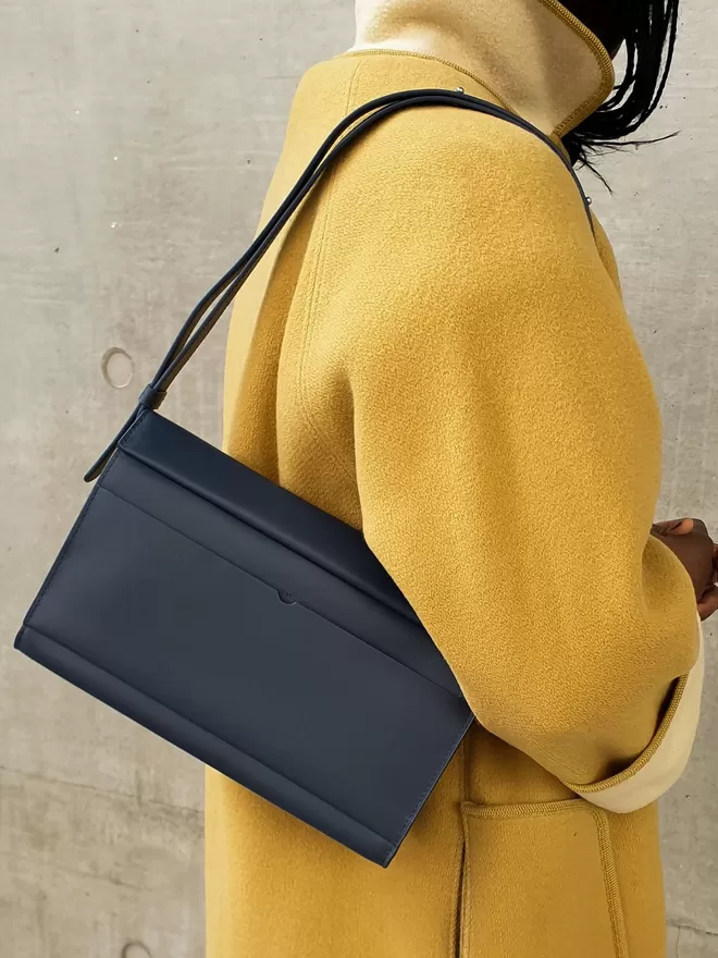 Navy shoulder bag adjusted to Shoulder Length and worn on an oversized amber wool coat. The model is facing the wall and has luscious braided hair.