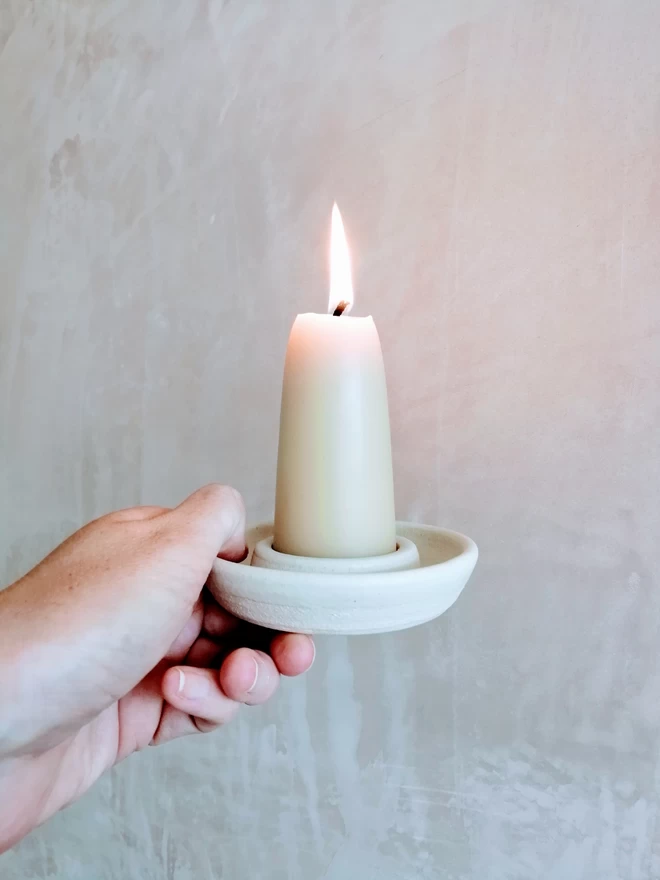Pearl white candle holder with lit short stubby beeswax candle. Held by a hand in front of bare plaster wall. candle creating soft glow