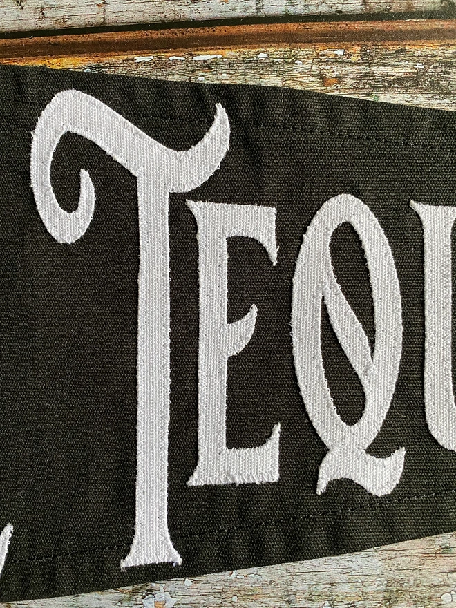 Detail of a black canvas Trick or Tequila pennant flag with the white canvas letters T E Q.