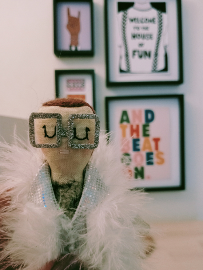 Close up of Elton John mini decorative icon doll showing his head and torso held in front of a small gallery of music themed prints Elton is wearing an all white faux leather feather trimmed outfit with oversized square framed silver glasses