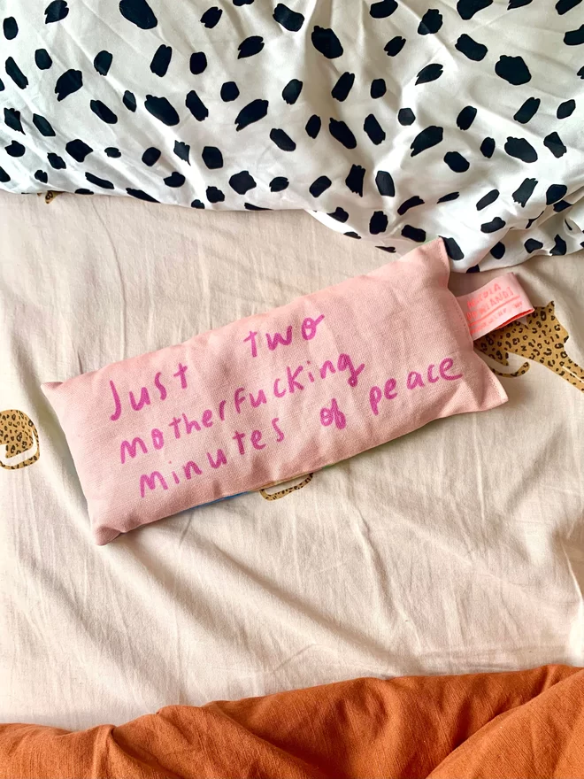 Lavender bag lies on bed near pillow. Peach fabric with magenta handwritten text 'Just two motherfucking minutes of peace'