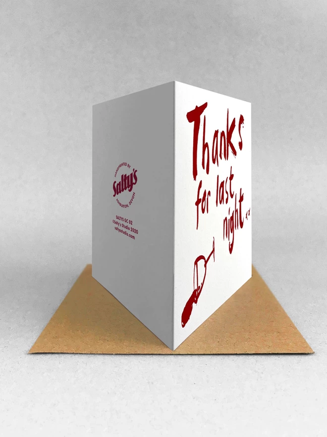 Rear view of a white greetings card with Thanks for Last night in a handwritten scrawl, screenprinted in claret red, with a knocked over wine glass besides. The card is stood on a brown kraft envelope in a light grey studio background.