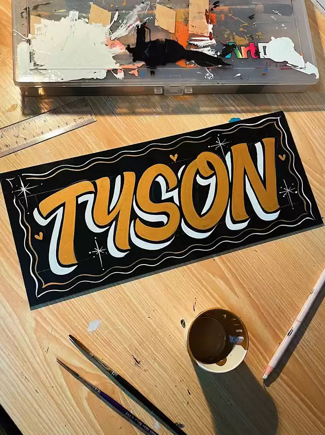Freshly hand-painted name sign spelling 'Tyson' with the materials used to create it around the piece, including paint brushes, ruler, a painty cup and wax pencil.