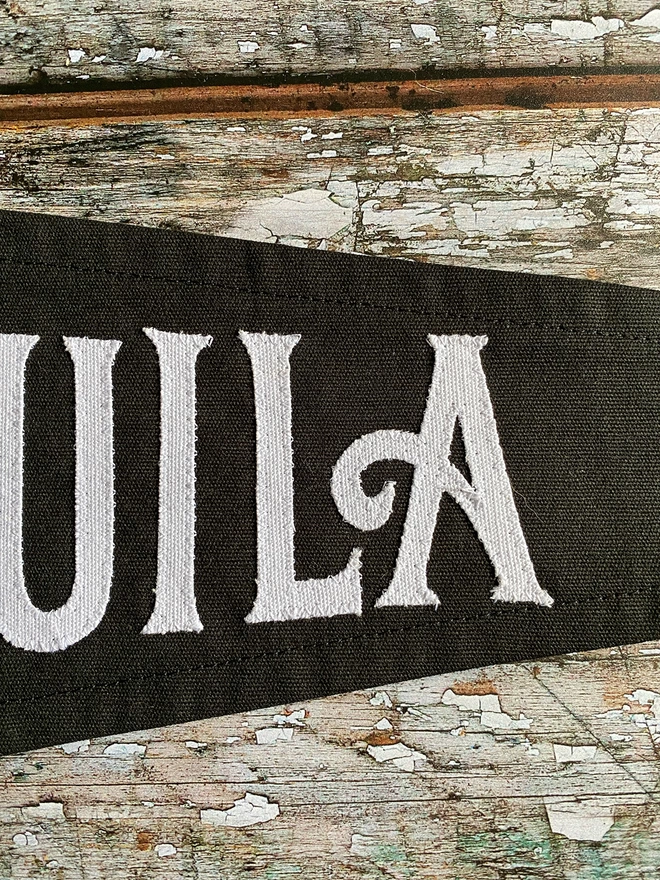 Detail of a black canvas Trick or Tequila pennant flag. You can see the letters U I L A in white canvas.