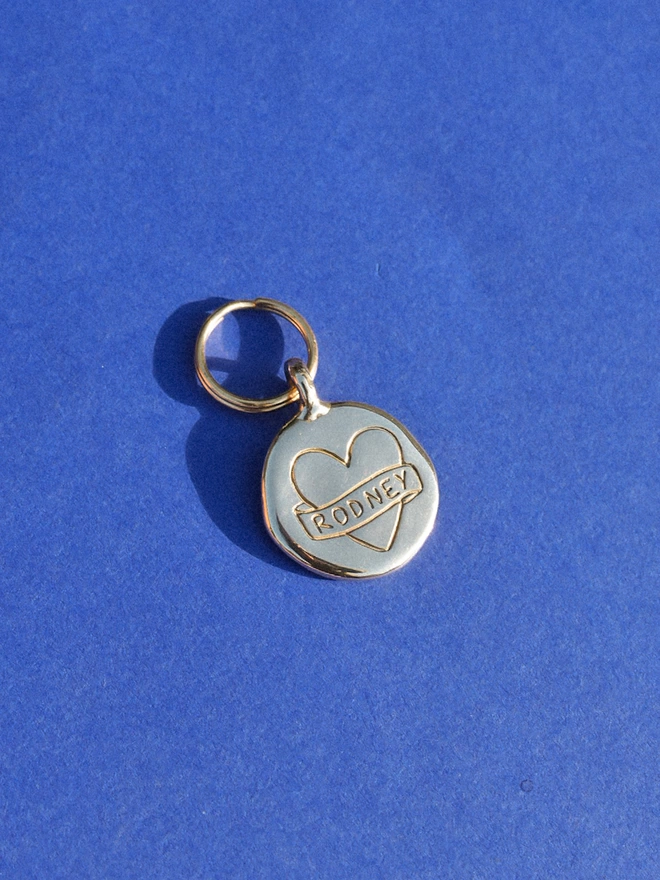 a shiny brass round pet tag with 'RODNEY' inside a banner wrapped around a heart, hand etched / engraved into the tag.
