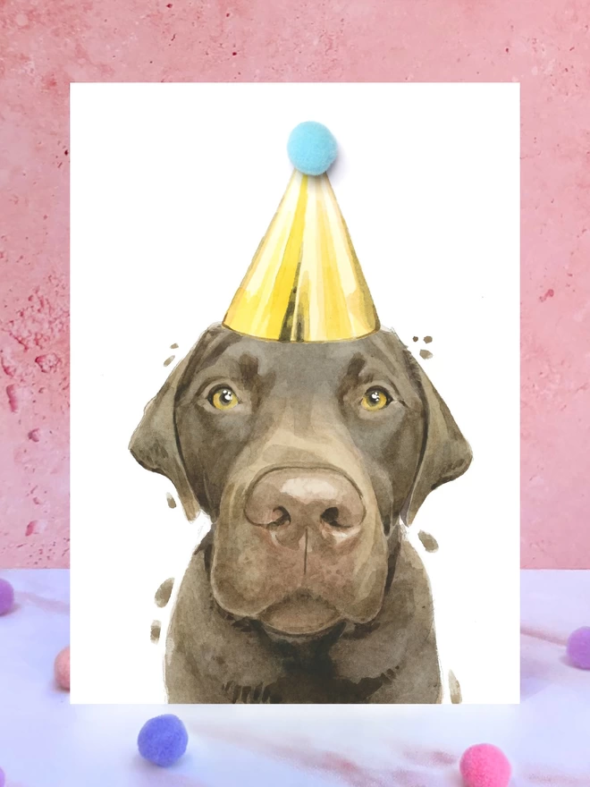 A greeting card featuring a hand painted design of a chocolate Labrador, stood upright on a marble surface surrounded by pompoms. 
