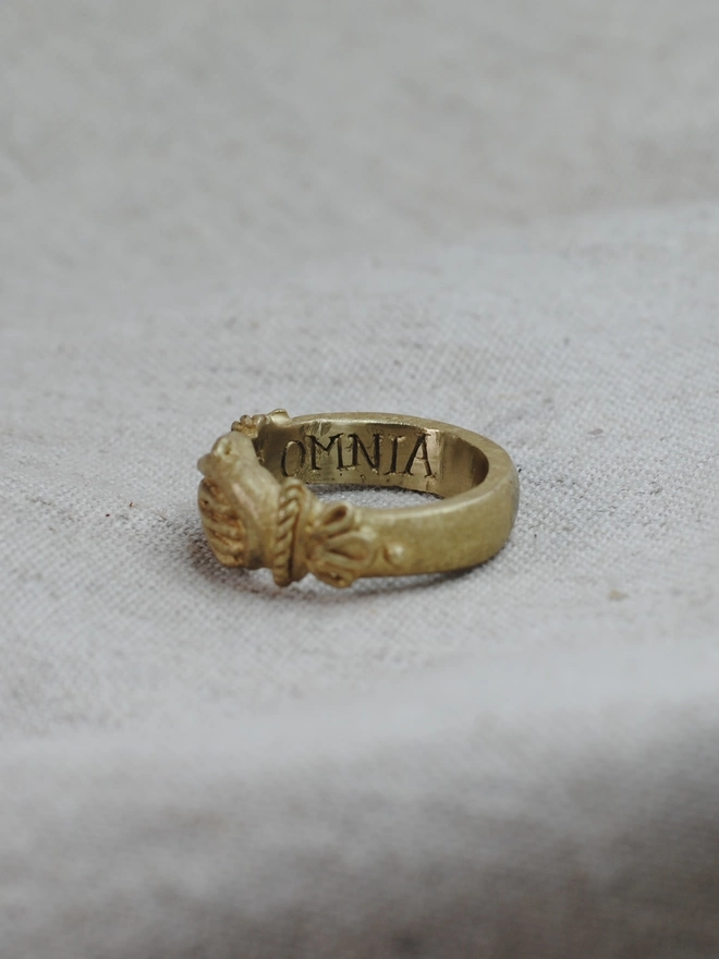 The image of a gold toned ring having being turned to the side to reveal the Latin word omnia engraved onto the inside of the band 