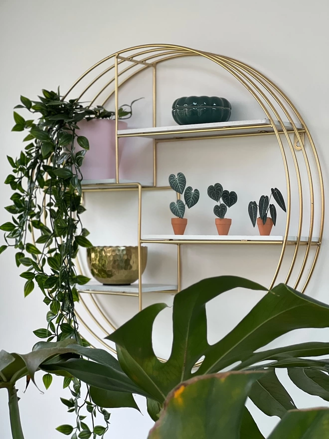 3 miniature paper plant ornaments on a gold round shelf system alongside other pink, green and gold ornaments with a real plant in the foreground