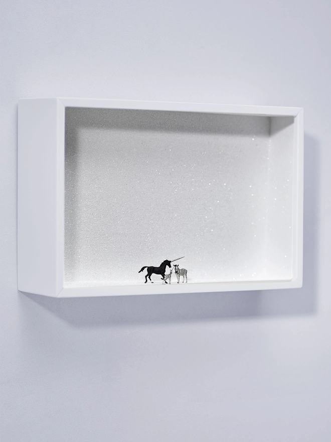 Miniature scene in an artbox showing a trio of tiny creatures - a black unicorn, a zebra and a baby ‘zunicorn’ (a mix of the zebra and unicorn) standing against a sparkling white backdrop 