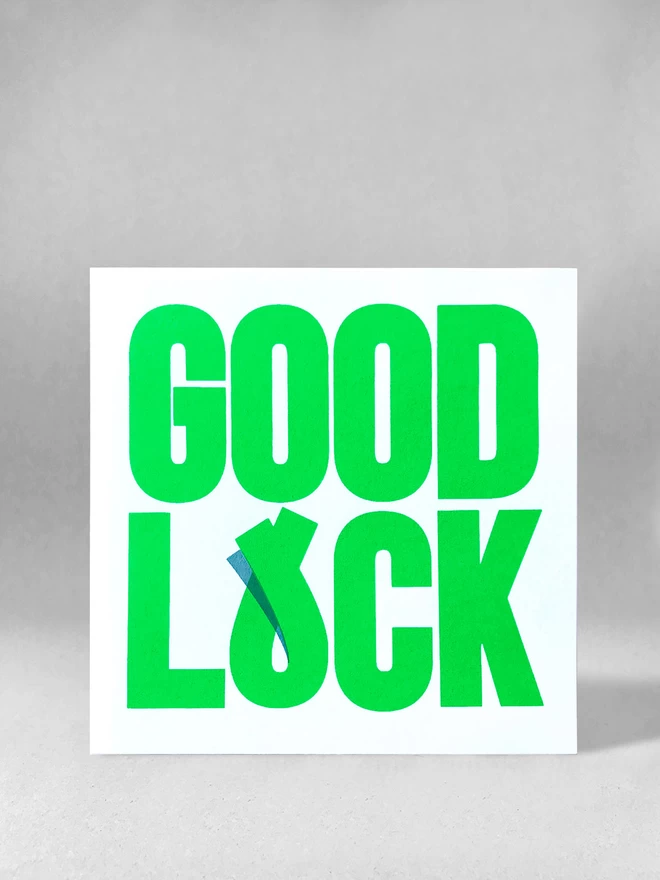 Green bold typography fills the card, ‘GOOD’ on the top, ‘LUCK’ on the bottom - the U has been tweaked to overlap suggesting fingers crossed for luck. There is a small shadow printed to make the overlap clear. This is a studio shot of the card stood front on to the camera, stood on a light grey background.