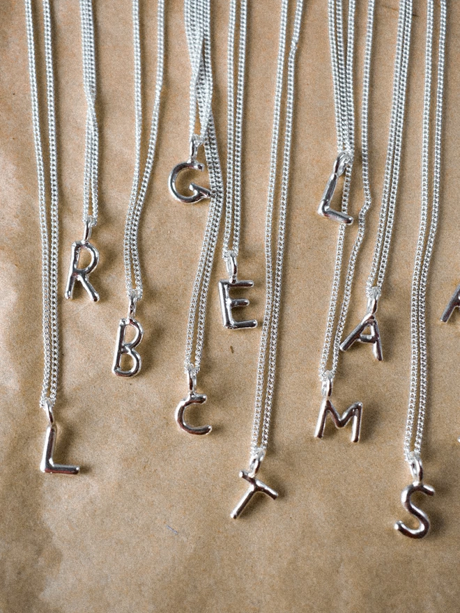 recycled silver alphabet pendants in a variety of letters, on thin silver chains, layered over each other on a brown paper background. 
