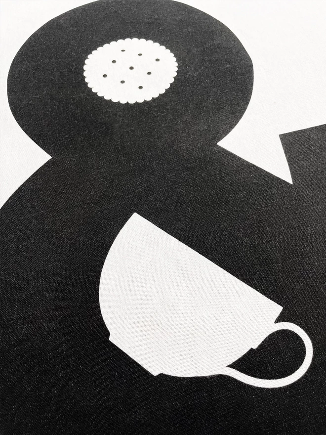 Close up of a tea towel showing the screenprinted black ink with a white cup shape and a biscuit within an ampersand.