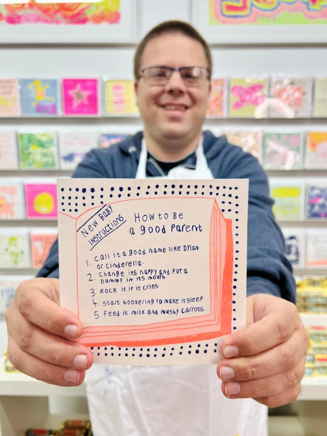 Artist holding A new baby card with a list of funny instructions printed in fluro orange & deep blue