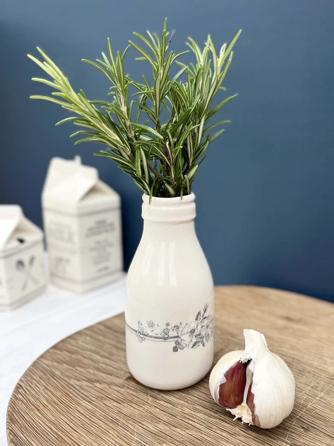A small handmade ceramic ‘milk’ bottle/vase is filled with a fresh bunch of herbs. 