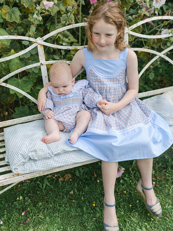 A girl holds a baby while sitting on a bench. They are both wearing matching checked clothes. the girl in a dress and the baby in a romper.