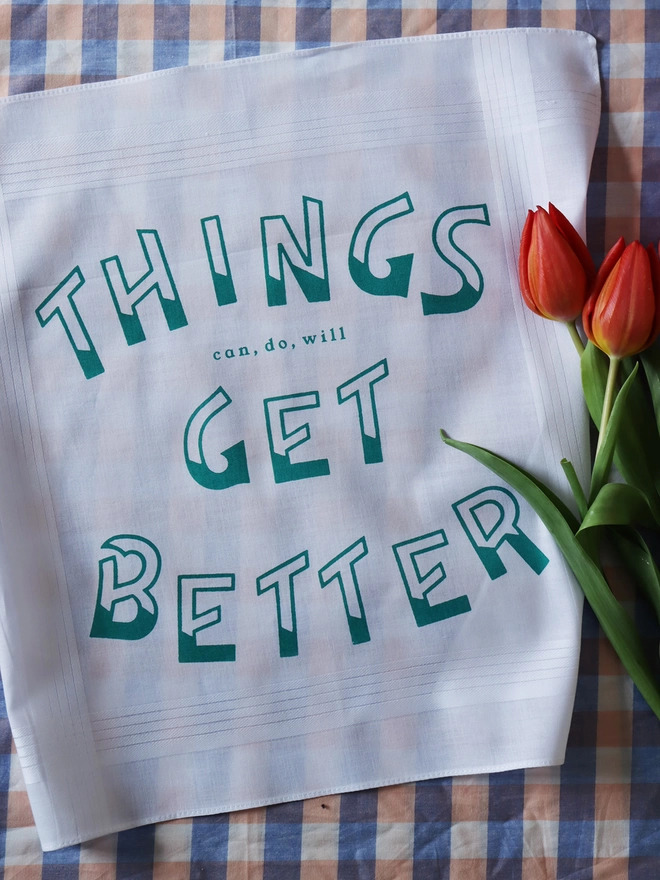 Mr.PS Things Get Better hankie printed in green laid on a gingham tablecloth alongside red tulips
