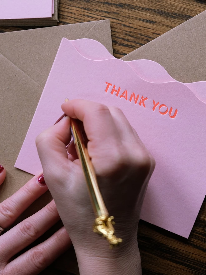 Lady writing on wavy pink card with 'Thank you' printed across the top in neon red ink.