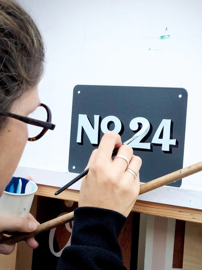 Sylvie painting a house number in flourish style, pale blue and black No. 24 on anthracite grey metal plaque.