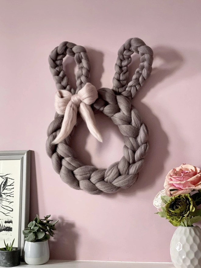 A soft grey merino rabbit wreath is hung on a light pink wall, it has a woolly bow to match.