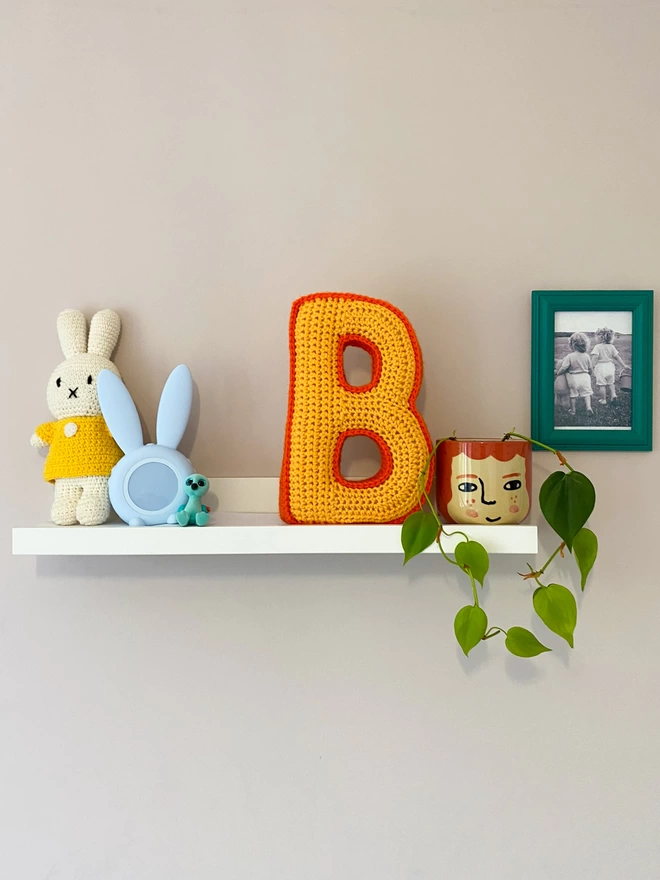 Crocheted cushion shaped like the letter B in Pale Orange and Orange on a shelf in a childs room