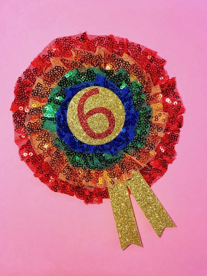A number 'six' rainbow sequin rosette. The centre is a gold circle with a red number 'six' in the middle. Going out from the centre are five layers of sequins in the colours blue, green, orange, and red. 