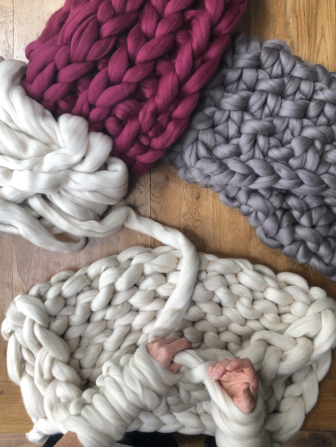 Taken looking down onto an oak table, image shows an oyster cream blanket being arm knitted, the big loops covering a white woman's forearms as she reaches for another stitch