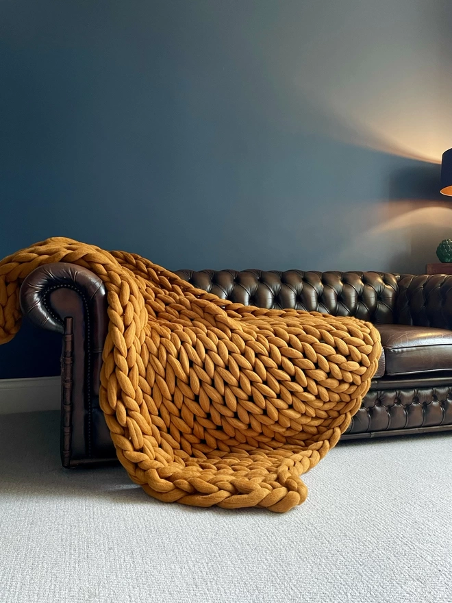 A rainbow merino arm knitted blanket cascading down the side of a brown leather Chesterfield sofa, before a dark blue wall