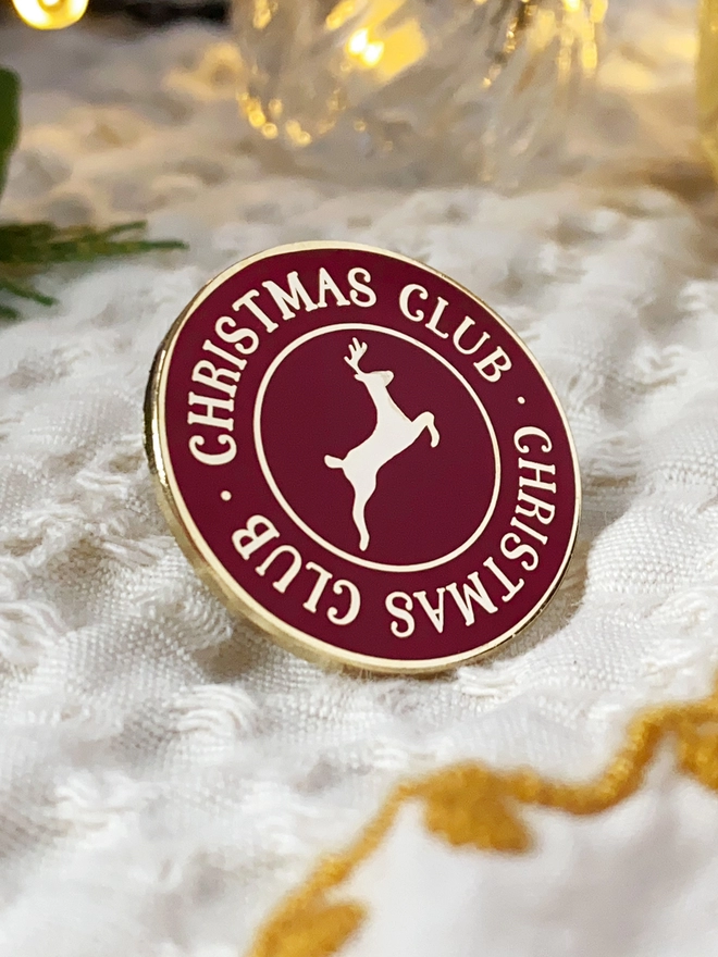 A deep red and gold enamel pin badge with a gold reindeer in the centre and the words "Christmas Club" around the outside is on a white blanket.
