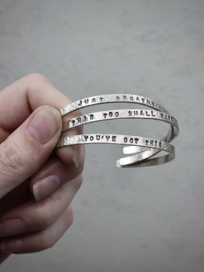 The left hand of a woman holding three sterling silver cuff bangles with hand-stamped text, above a pale green background.