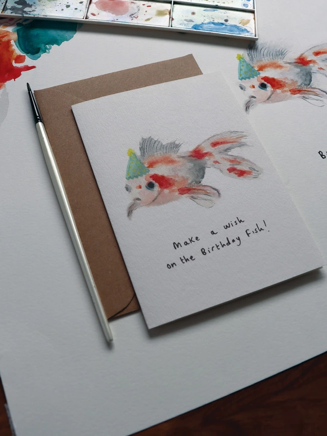 Close Up Slightly Out Of Focus Shot Of The Goldfish Greeting Card Sitting Along Side The Original Hand Painted Watercolour Illustration, Paintbrush and Palette