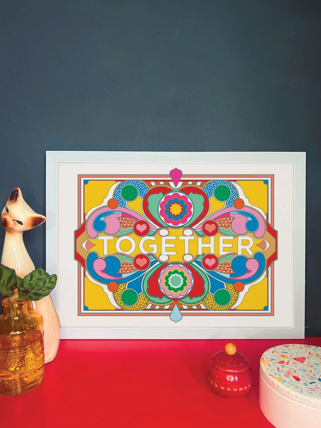 A landscape print on a white background with dark blue linework and green, blue red pink and gold sections. Across the centre of the image is written ‘TOGETHER’. The print is in a white frame on a red cabinet, and is resting against a dark grey wall. Next to the frame is a cat ornament, an orange vase with a plant, a small red wooden pot and a white and multi coloured terrazzo pot.