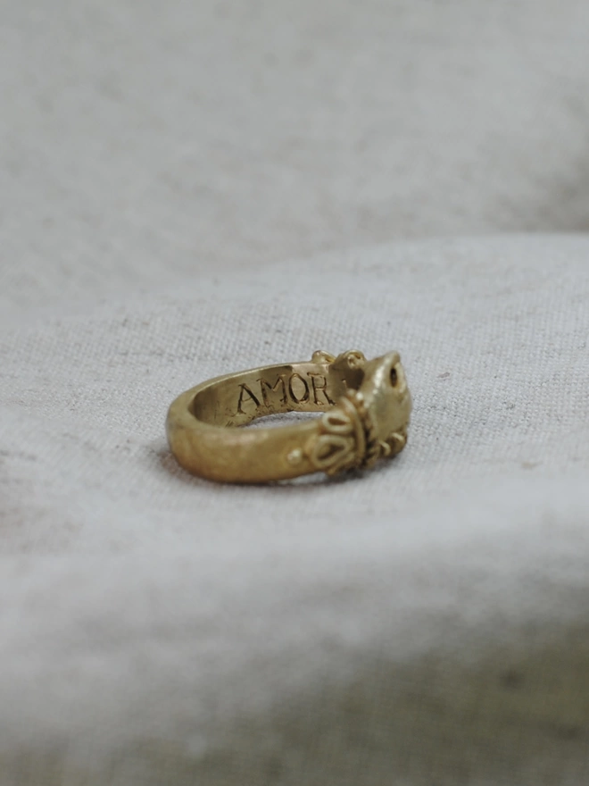 The image of a gold toned ring having being turned to the side to reveal the Latin word Amore engraved onto the inside of the band 