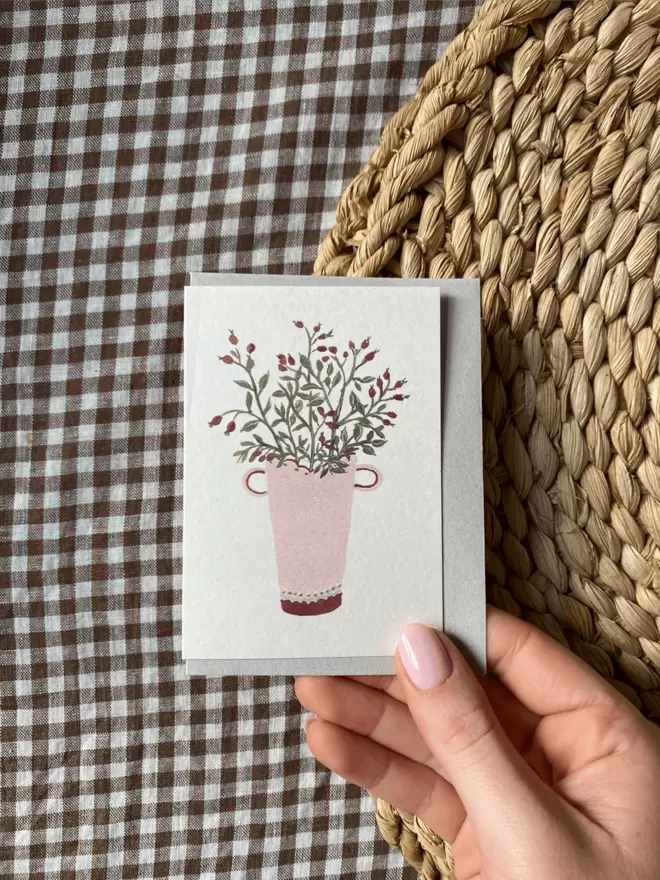 mini greetings card with rosehips on in a vase, over brown gingham tablecloth.