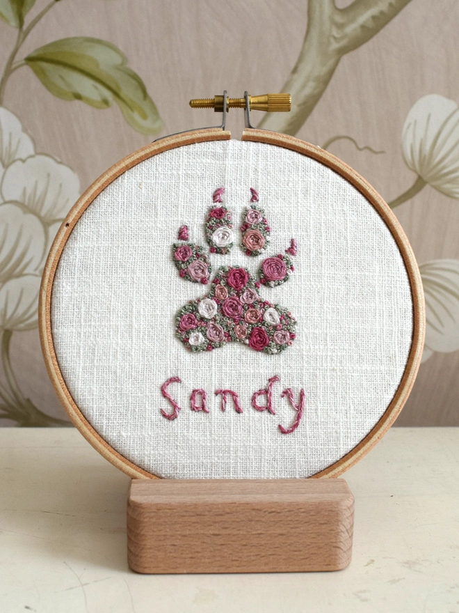 Pink Roses Dog Paw, an embroidery design of Woven Roses and scattered French Knot blossoms in 5 shades of pink with 2 shades of green grass French Knots.  Displayed in an embroidery hoop on a wooden stand.