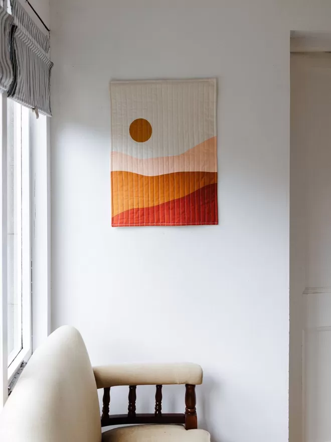Earth Quilt Hanging On White Wall Above Cream Chair