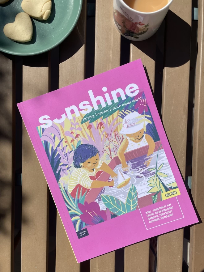 Sonshine Magazine Issue 20 , a pink magazine with an illustrted image of two children playing in water together.. The magazein is lying on a marble table top with a cup of tea and heart shaped biscuits