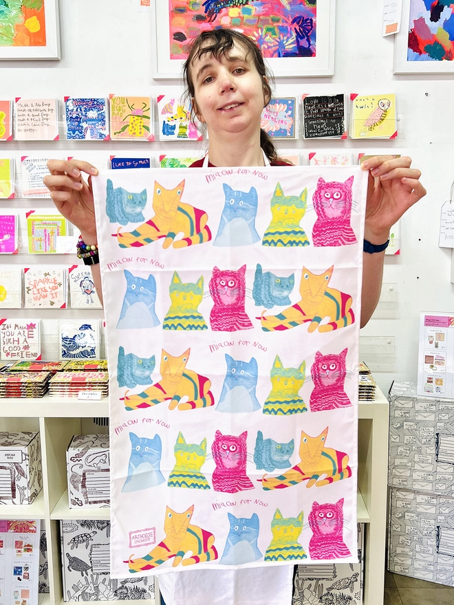 Happy artist holding miaow for now 100% organic cotton charity tea towel in blue pink & yellow